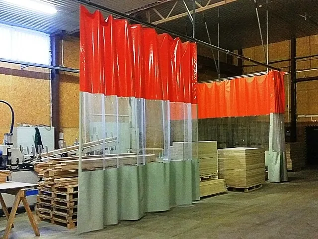 Industriāls PVC aizkars ar oranžu augšdaļu un caurskatāmu joslu, kas sadala kokapstrādes darbnīcas zonas